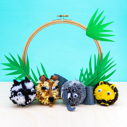 Pom-pom animals attached to a wooden embroidery hoop with green leaves on a blue background
