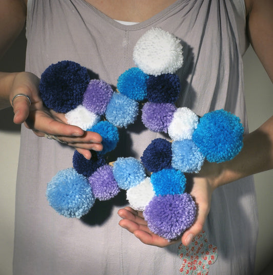 Person holding a colorful pom-pom wreath made of blue, purple, and white pom-poms.