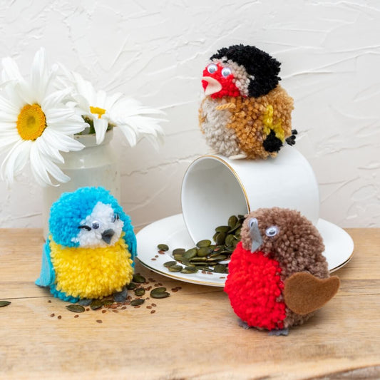Three colorful knitted bird toys on a table with a cup and saucer.