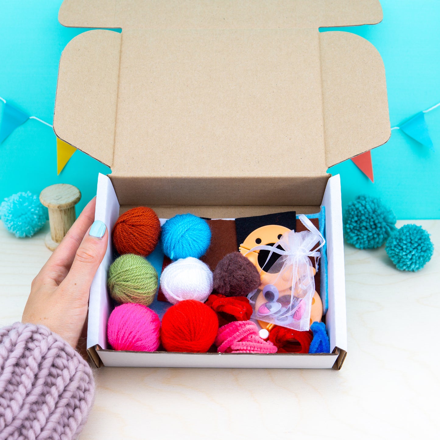 Open cardboard box with colorful yarn balls and a small plush toy on a light blue background.