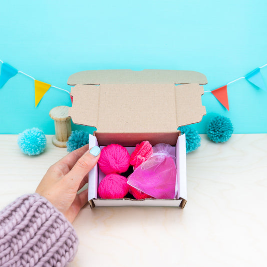 Hand holding a box of pink yarn against a blue background with decorative flags.
