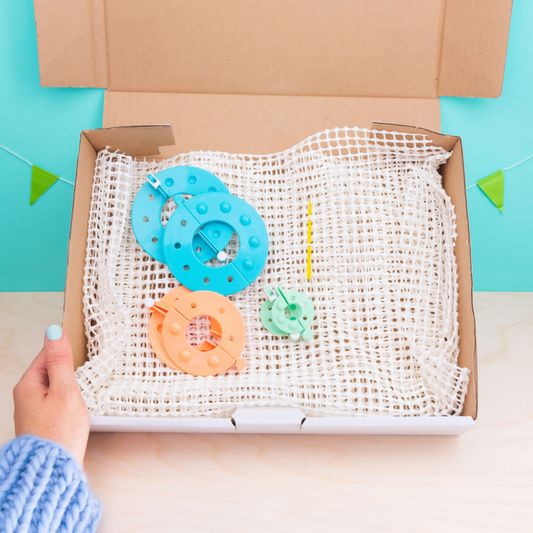 Open cardboard box with craft materials including a blue wheel, orange flower, and green leaf on a blue background.