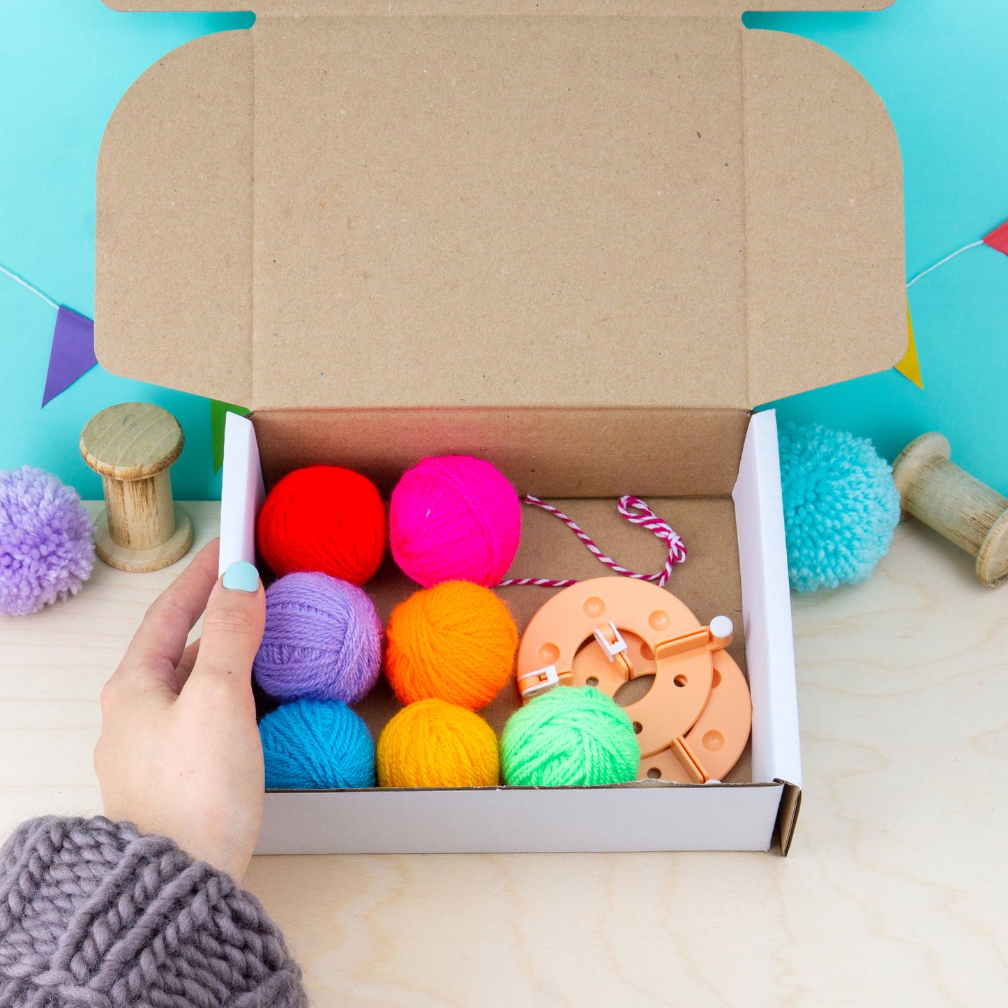 Box of colorful yarn balls and knitting tools on a wooden surface with a hand holding a crochet hook.