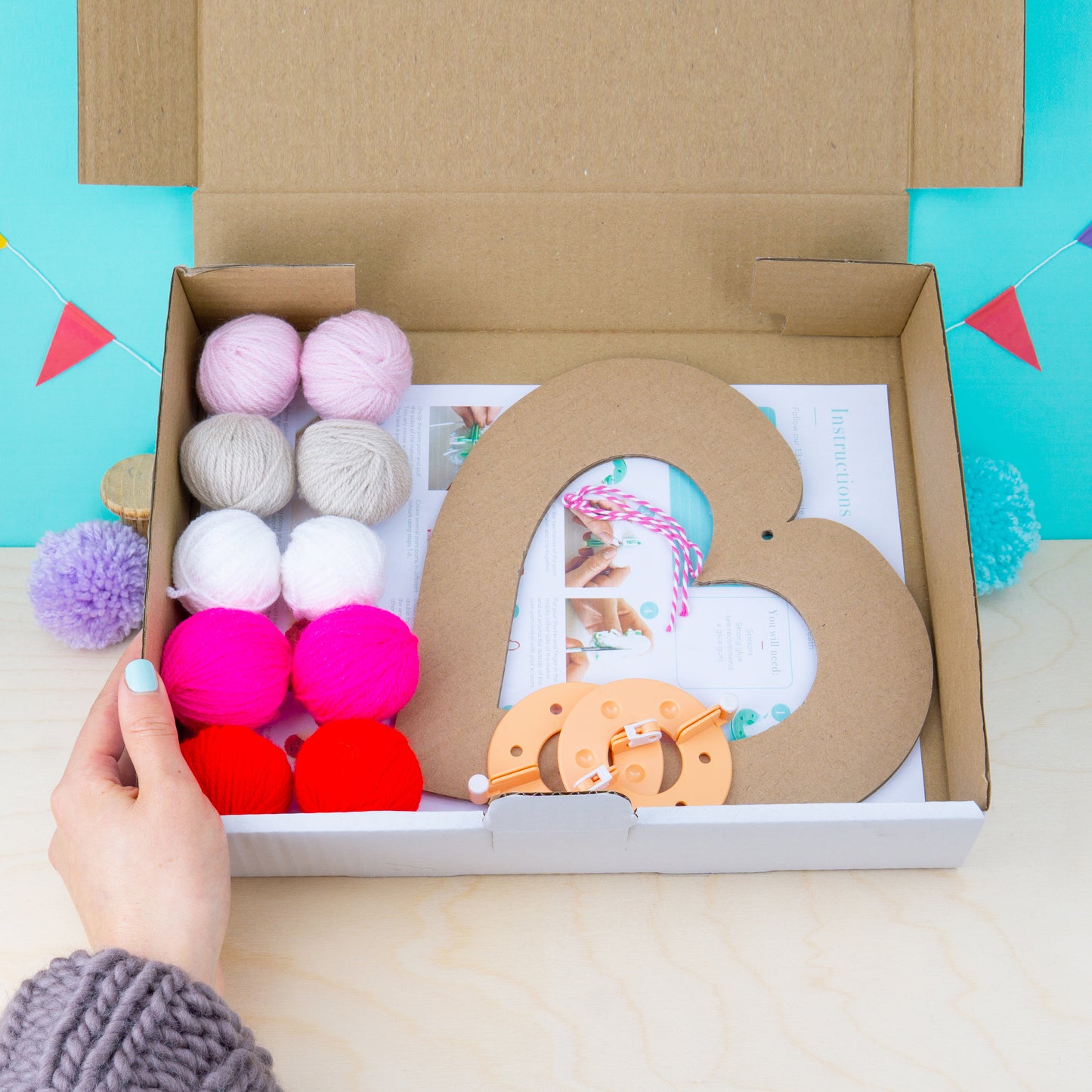 Cardboard heart-shaped frame with colorful pom-poms and a small toy in an open box against a blue background.