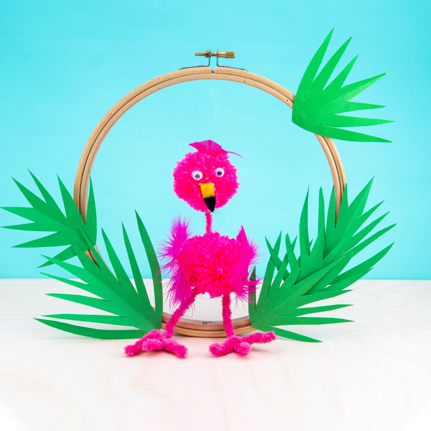 Pink flamingo craft made from pom-poms on a wooden hoop with green leaves against a blue background