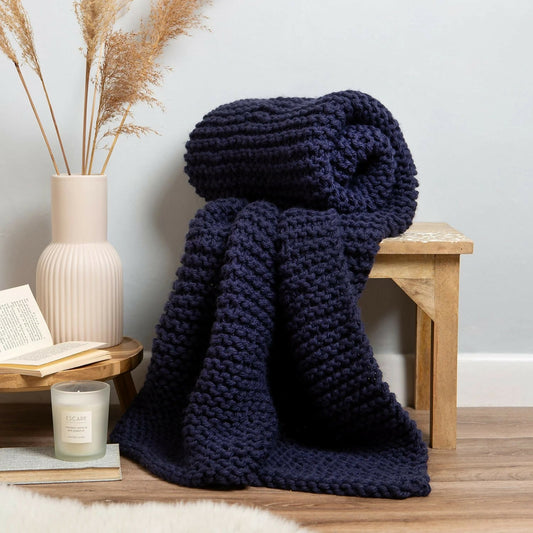 Navy blue knitted blanket draped over a wooden stool with a candle and book on a side table.