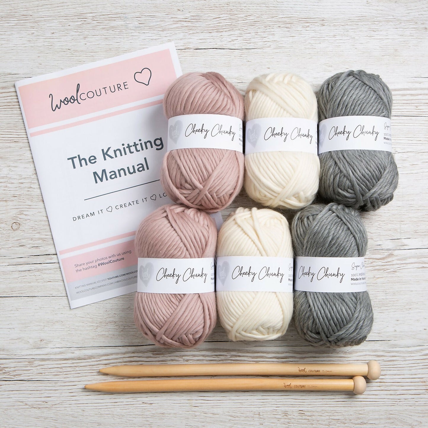 Set of yarn balls in pink, white, and gray with knitting needles and a manual on a wooden surface.