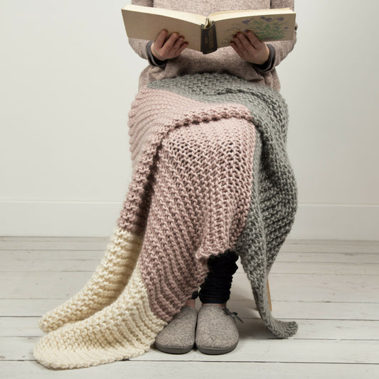 Person sitting on a wooden floor with a knitted blanket draped over their legs, reading a book.