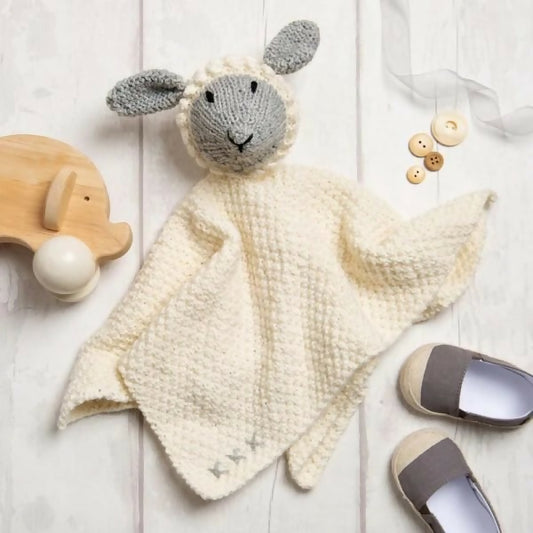 Knitted sheep comfort blanket on a light wooden surface with toys and shoes.