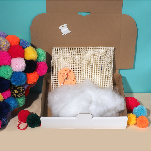 Open cardboard box with craft materials including pom-poms and a woven frame on a teal background.