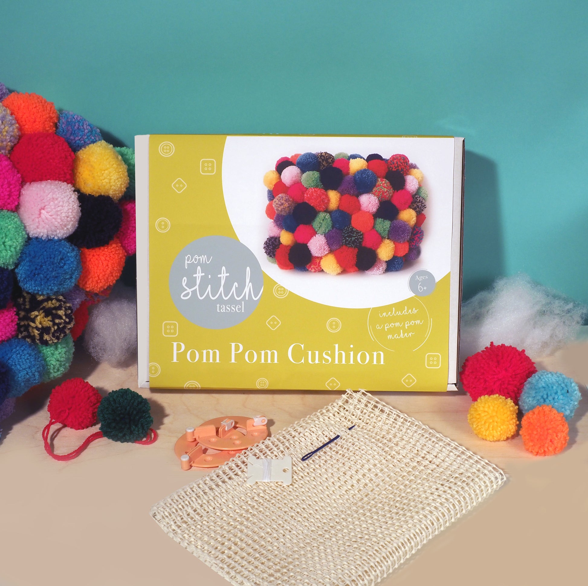 Pom pom cushion kit with colorful pom poms, a crochet hook, and yarn on a teal background.