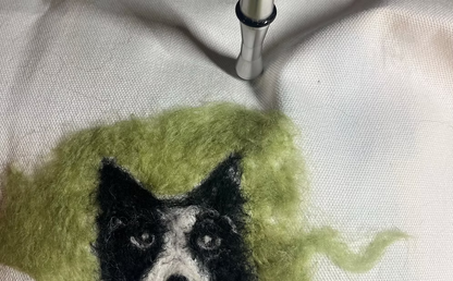 Felted wool art of a dog's face with green fur on a textured surface