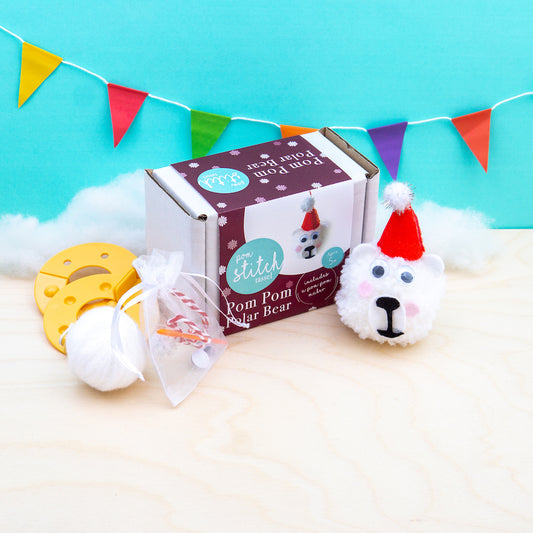 Pom pom craft kit with a completed pom pom polar bear, packaging, and tools on a light blue background with colorful flags.