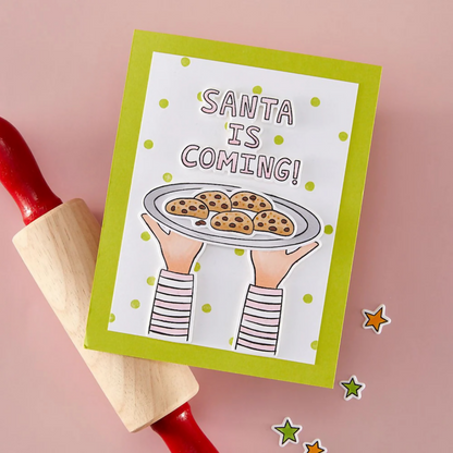 Card with 'Santa is Coming!' text and a plate of cookies, held by hands, on a pink background with a rolling pin.
