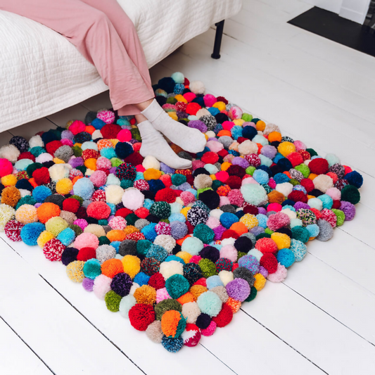 Colorful pom-pom rug on a white floor with a person wearing pink pants and white socks.