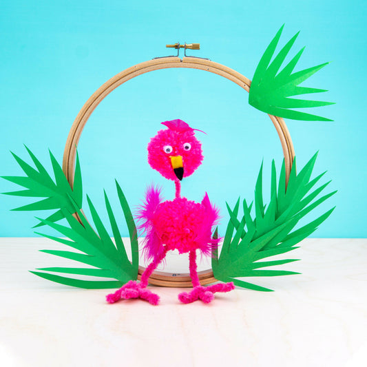 Pink flamingo craft made from pom-poms on a wooden hoop with green leaves against a blue background
