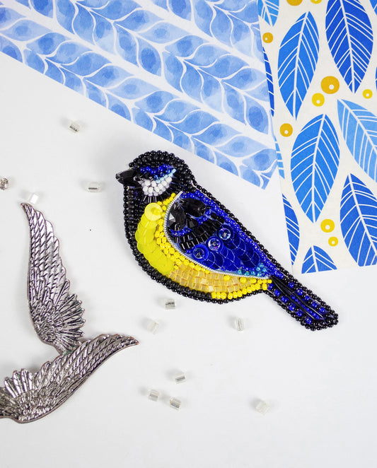 Decorative bird brooch with blue and yellow colors on a white background with leaf patterns.