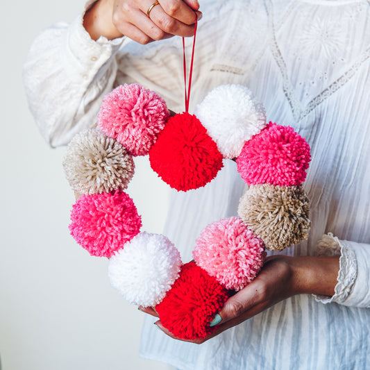Heart-shaped wreath made of colorful pom-poms held by a person against a light background