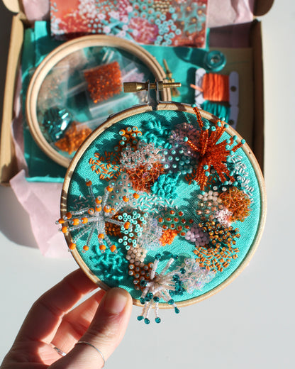 Embroidery hoop with beadwork held by a hand, with a box of embroidery supplies in the background.